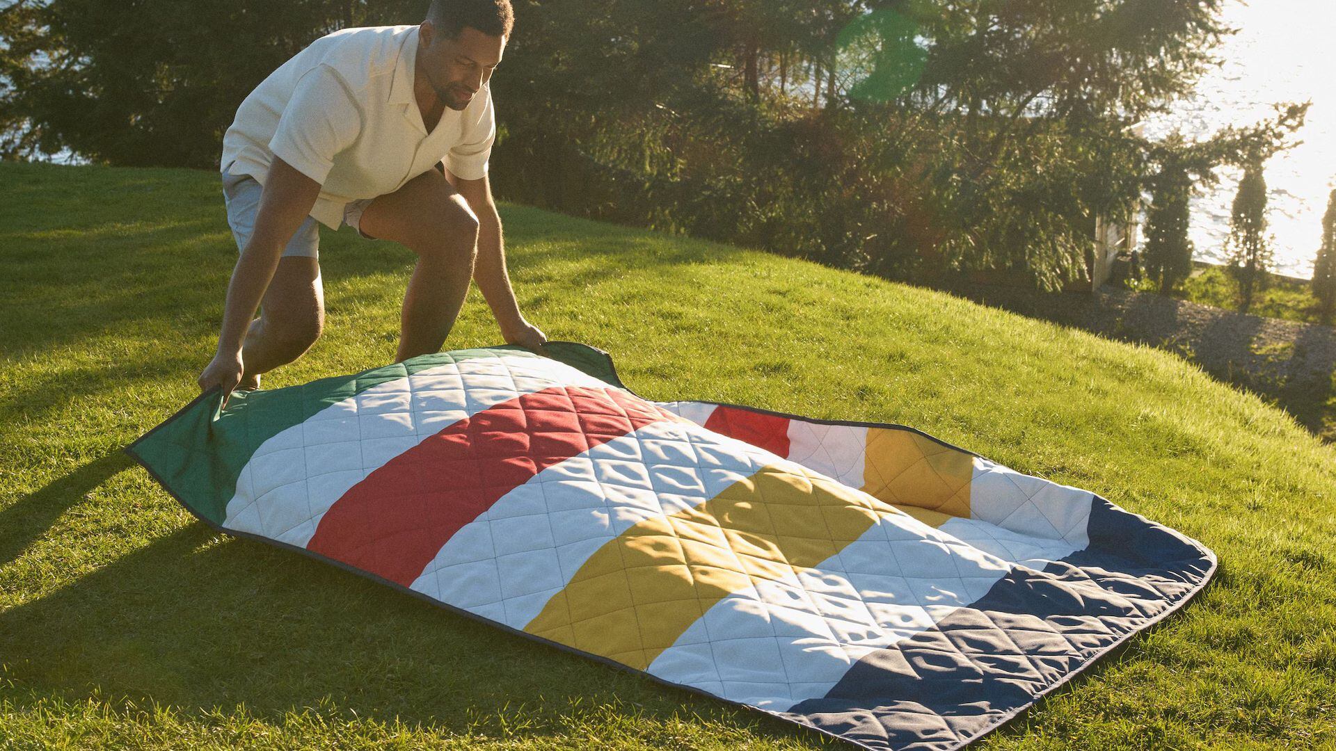 A person laying out a Canadian Tire Hudson's Bay striped blanket on a grassy lawn.