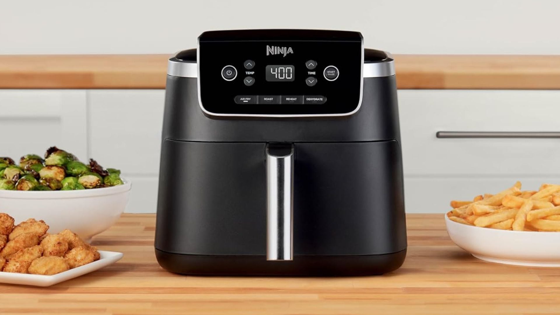 A Ninja Air Fryer Pro on a kitchen counter surrounded by Brussels sprouts, chicken wings, and French fries.