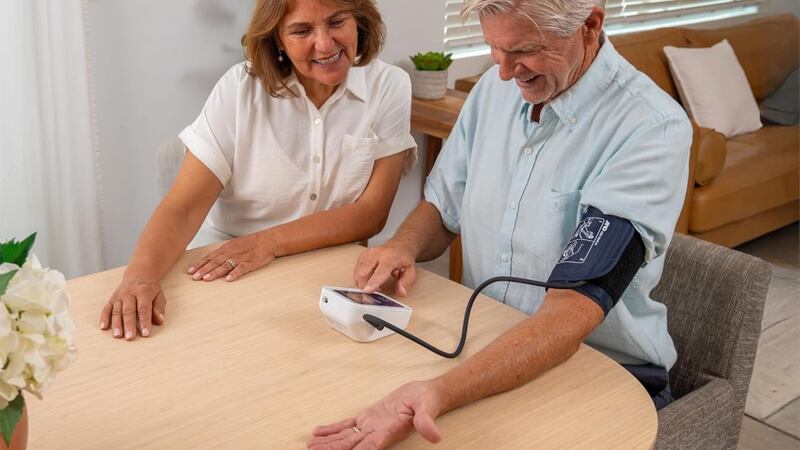 Two people are sitting at a table, one is using a blood pressure monitor.