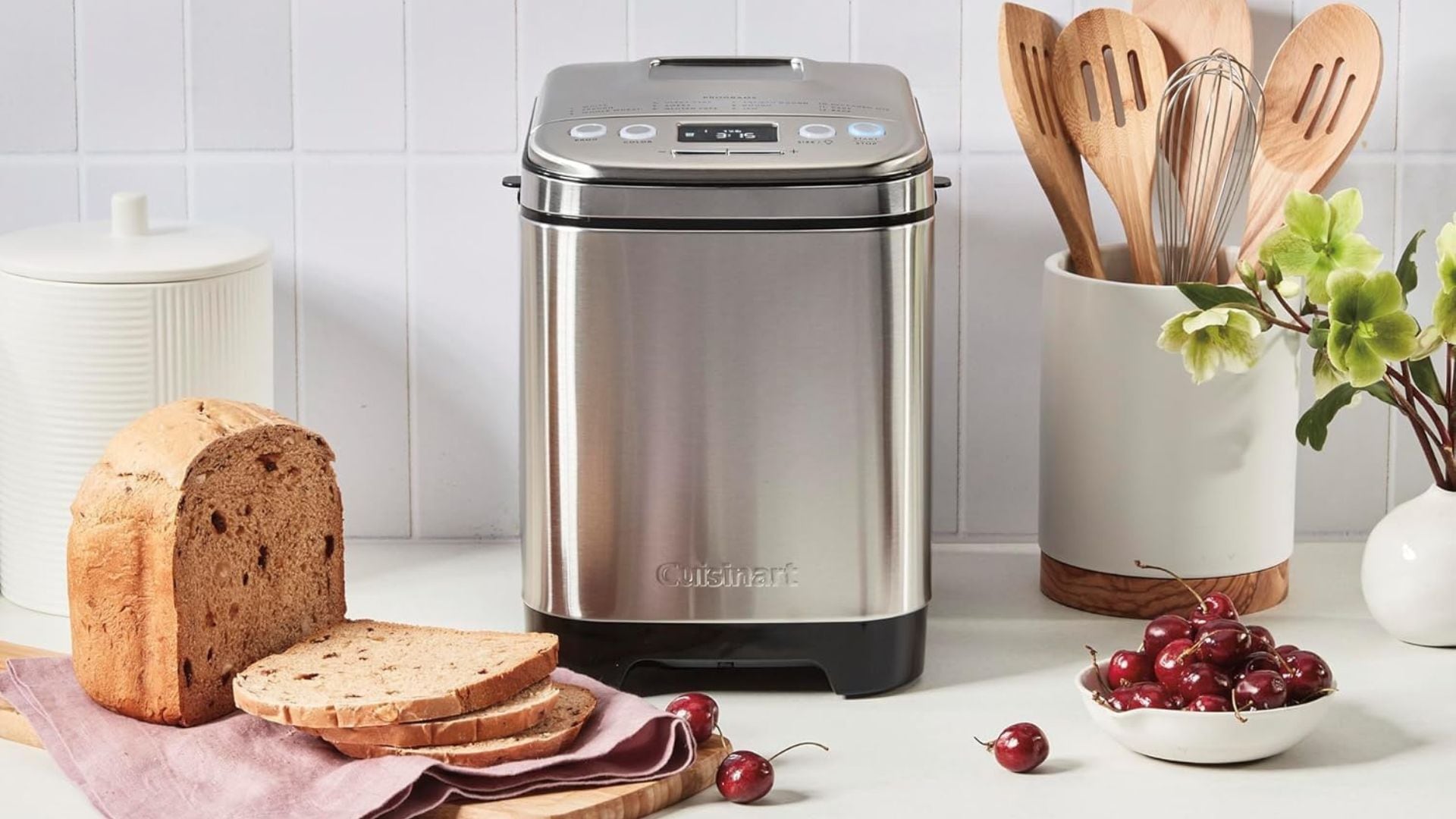 A Cuisinart Bread Maker on a counter surrounded by utensils, cherries, and bread.