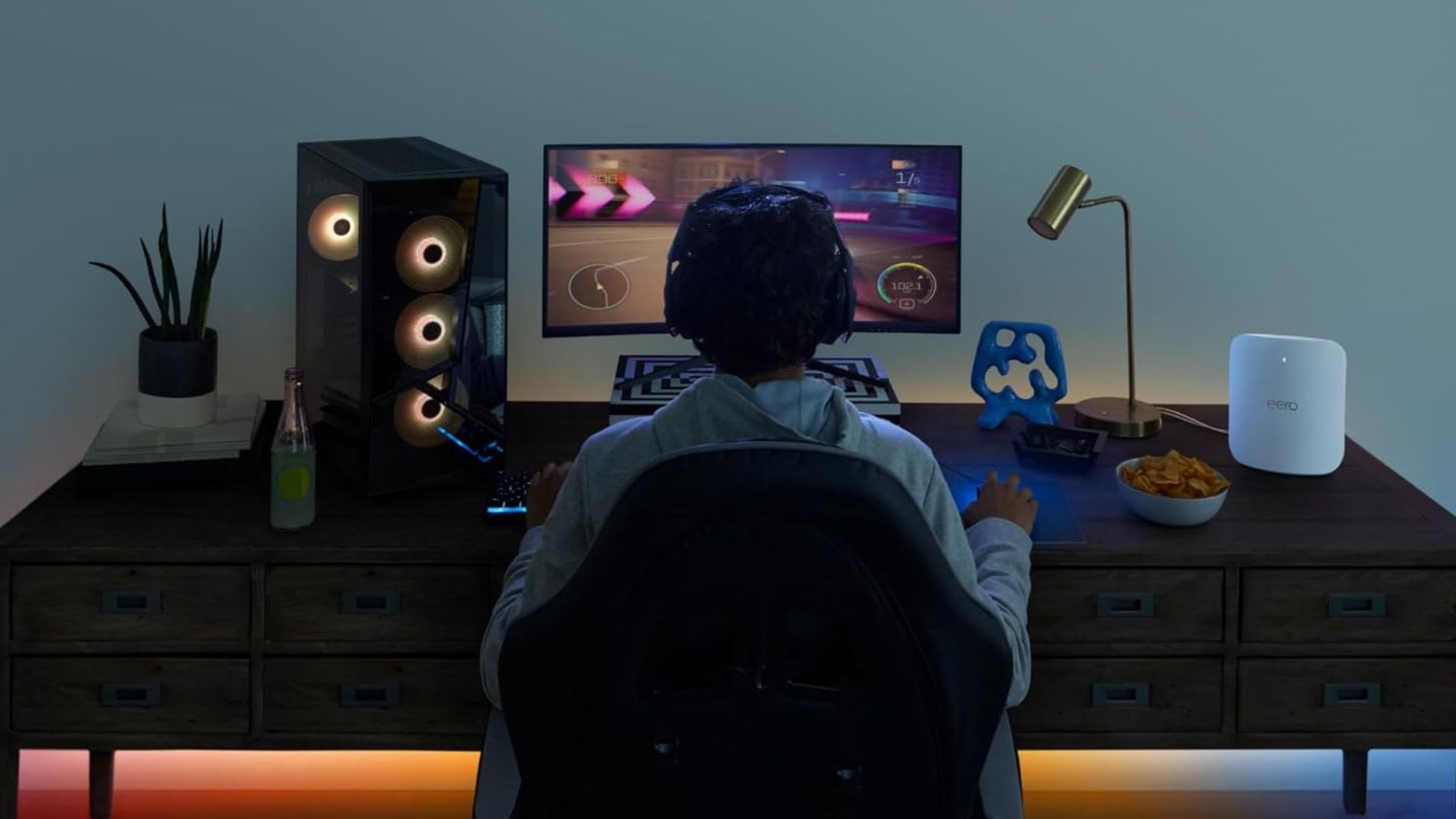 A person playing video games at a desk with the mesh router beside their computer.