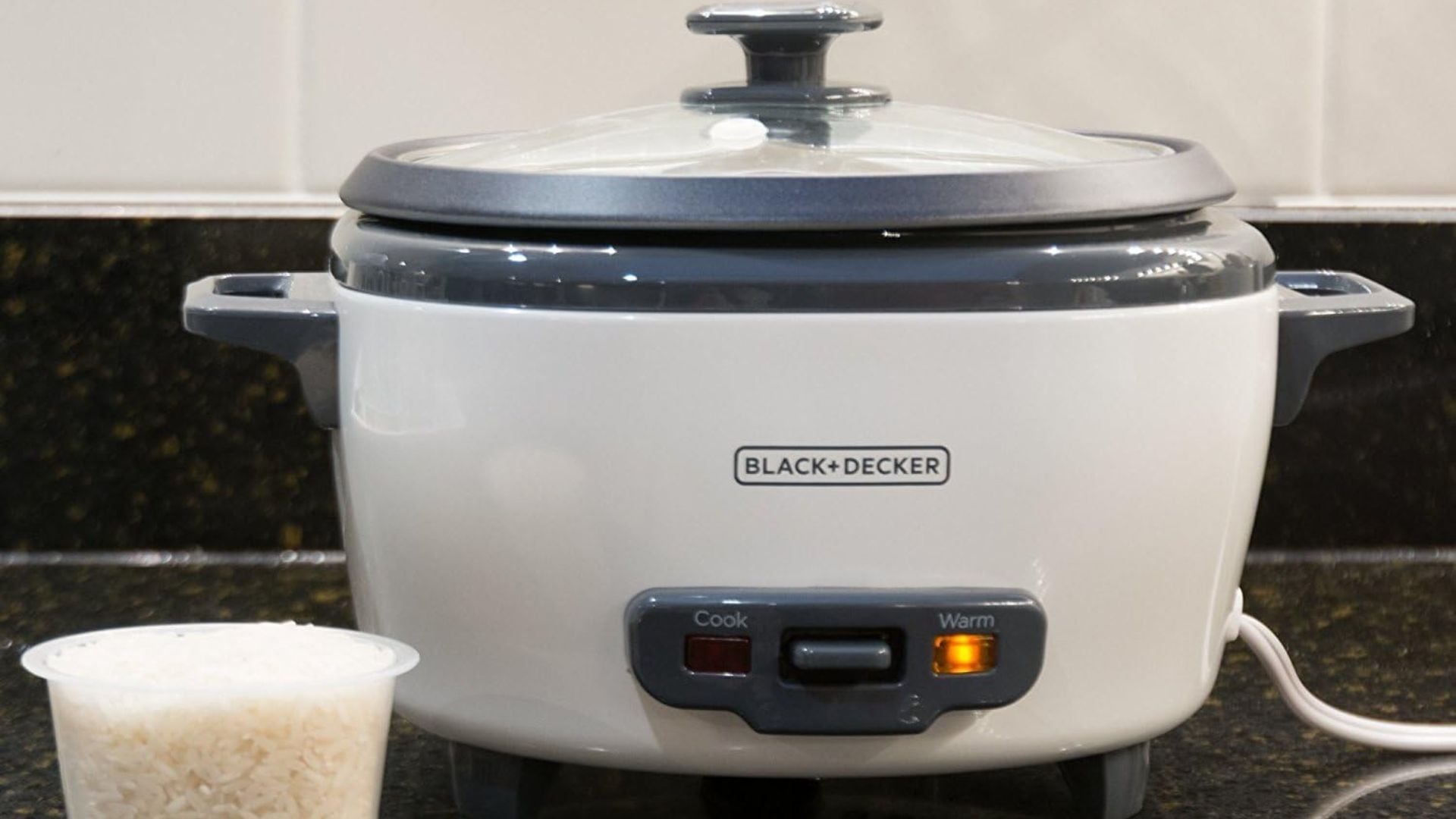 A rice cooker on a counter with a cup of rice next to it.