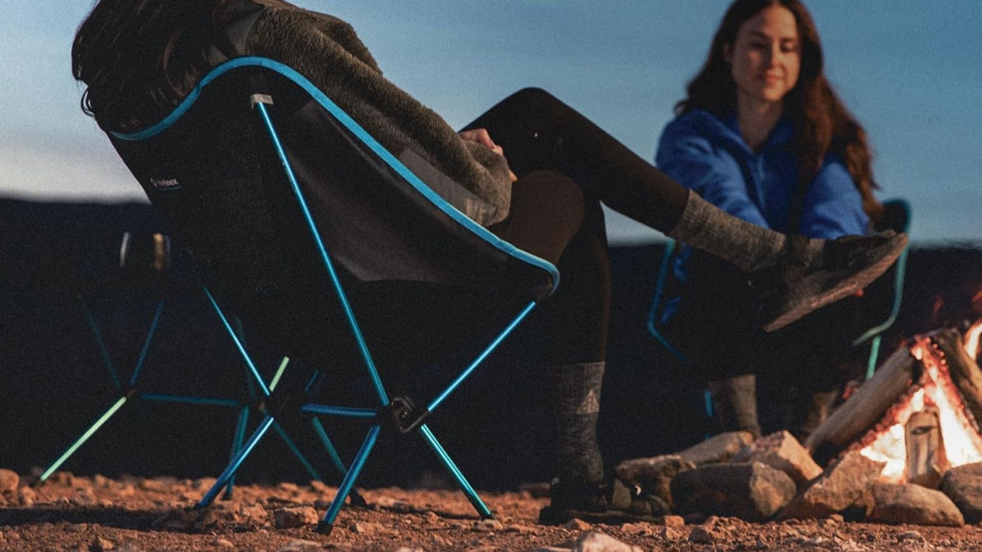 Two people sitting by a campfire using Helinox chairs.
